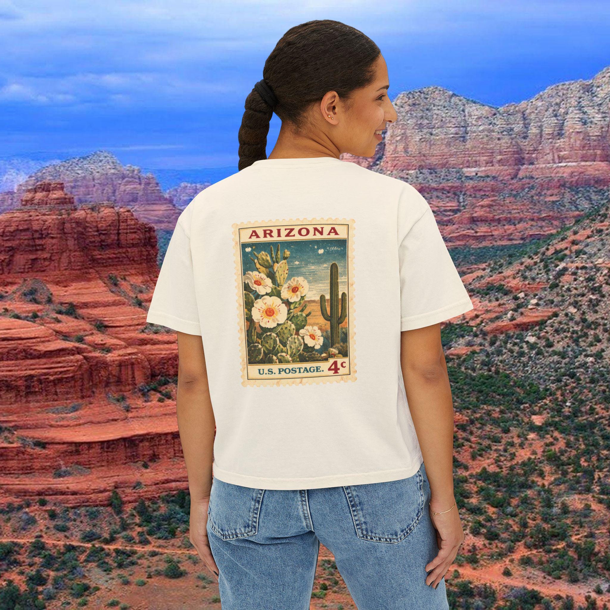 May include: Cream-colored t-shirt featuring a vintage Arizona postage stamp design. The stamp depicts a cactus, flowers, and the text "ARIZONA" and "U.S. POSTAGE 4¢". The shirt is paired with blue jeans, set against a scenic desert backdrop.
