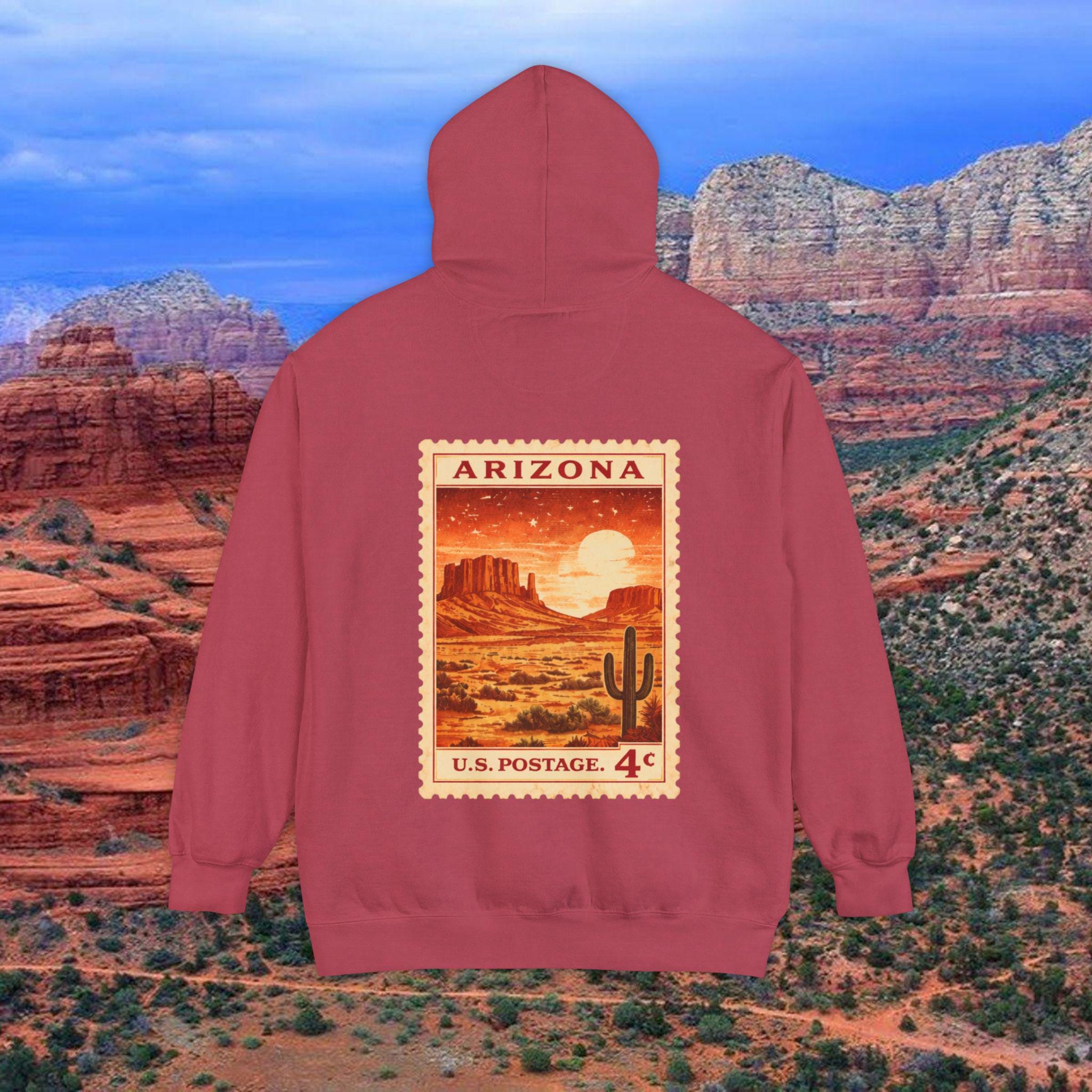 May include: A maroon hoodie featuring a vintage Arizona postage stamp design. The stamp depicts a desert landscape with a cactus, mesas, and a setting sun. The text on the stamp reads "ARIZONA" and "U.S. POSTAGE. 4¢".